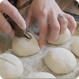 Closeup of hands using a scoring tool to score sourdough rolls