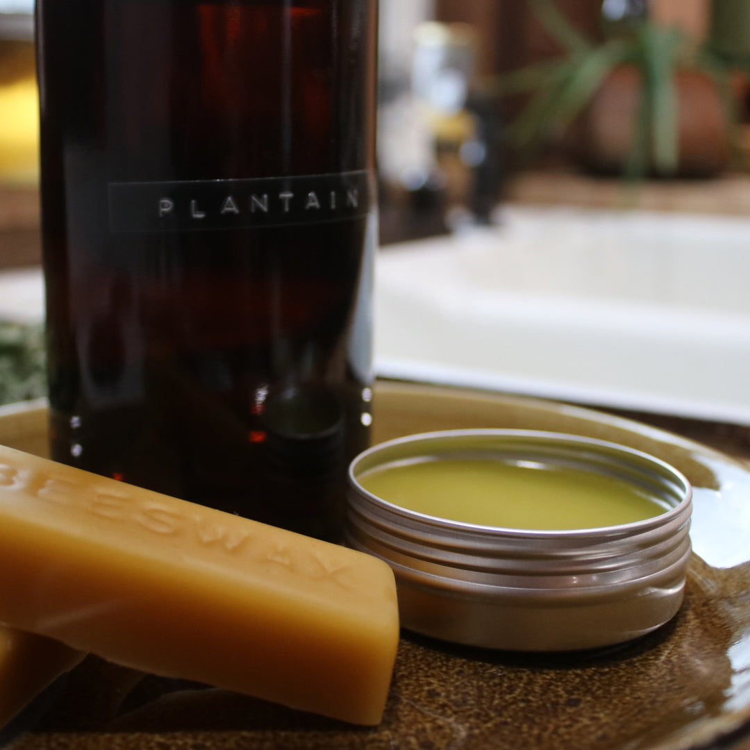 Close-up of natural skincare products including a dark "PLANTAIN" bottle, yellow balm tin, and bar of soap on a wooden surface.