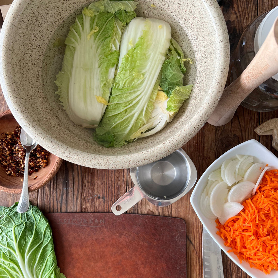 Kitchen prep scene with napa cabbage in speckled ceramic bowl, wooden cutting board, measuring cup, and plate of sliced onions and shredded carrots for fermentation.