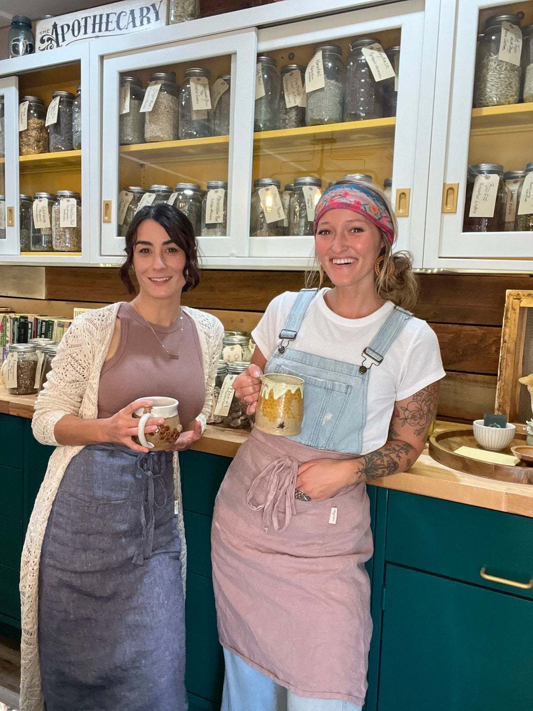 Jill and Kaylee in aprons holding mugs in a store with shelves of jars.