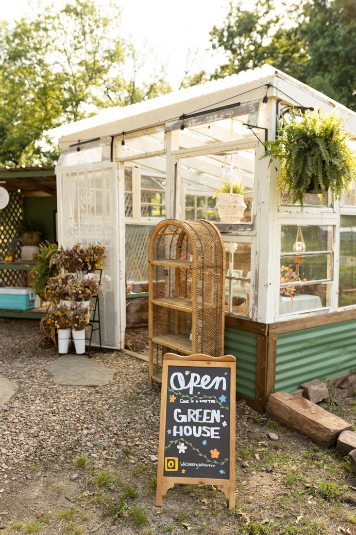 The white-windowed greenhouse shop with an Open sign, a fern, and dried flower buckets out front