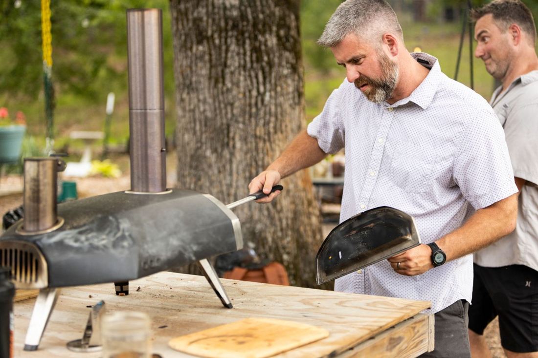 A man checking the pizza oven with a turning peel while a friend looks on