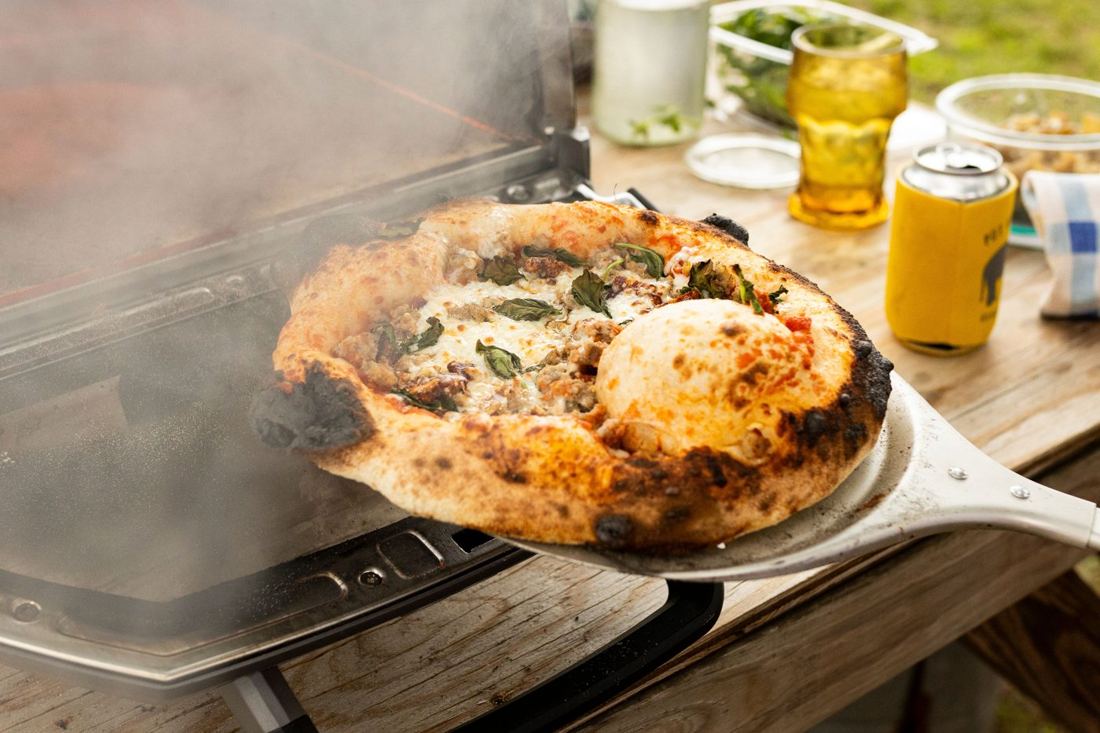 A blistered pizza with sausage, basil and mozzarella coming out of the steaming oven on a peel