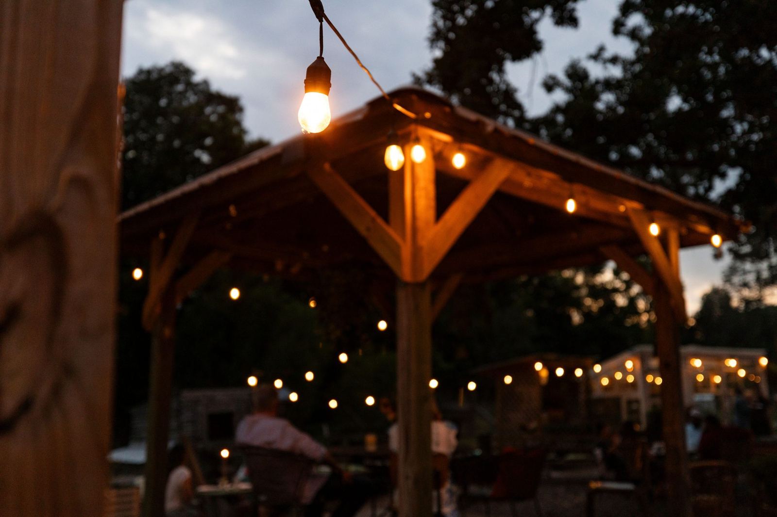 String lights glowing over a wooden pavilion and guests gathered in the garden at dusk