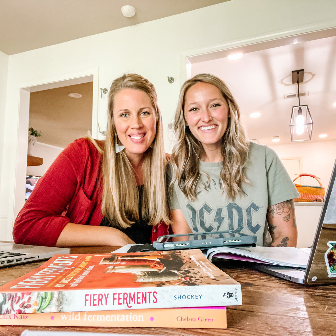 Jill and friend sitting at wooden table with laptops and fermentation books including "Fiery Ferments" and "Wild Fermentation" visible.