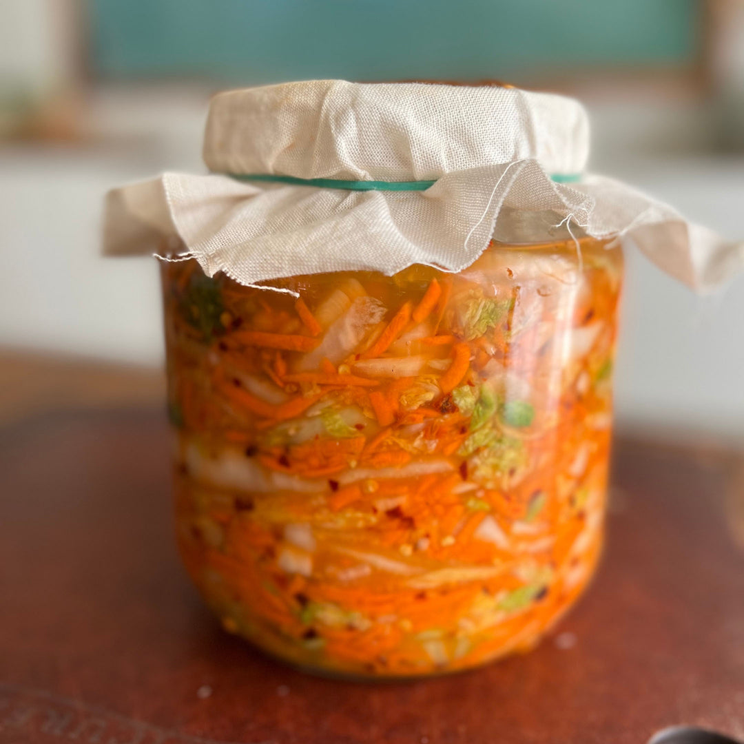 Glass jar with cheesecloth cover containing shredded carrots and napa cabbage, fermenting on a kitchen counter.