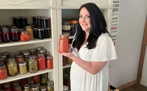 Woman holding a jar of red sauce in front of shelves filled with various jars.