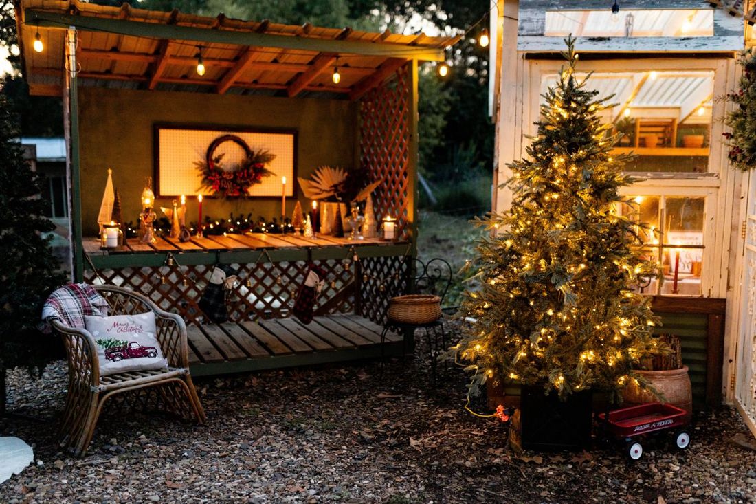 The candlelit potting bench with stockings and a wicker chair with a plaid blanket next to a lit tree