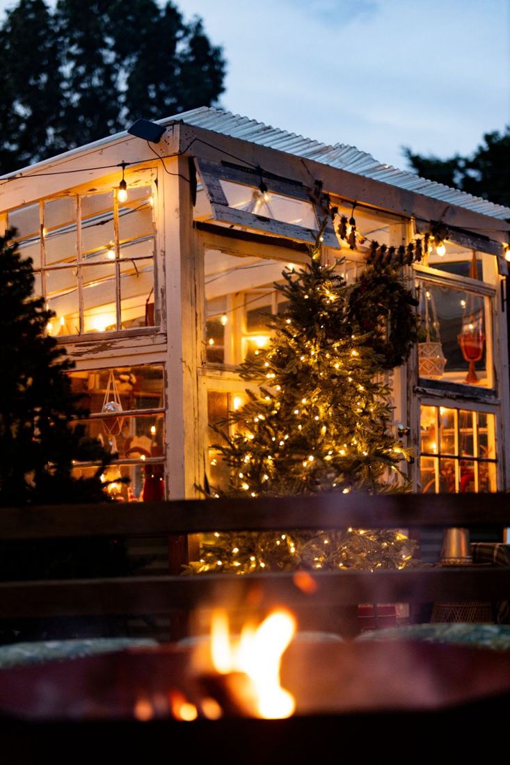 The white-windowed greenhouse glowing at dusk with a lit Christmas tree and wreath, a fire pit flickering in the foreground